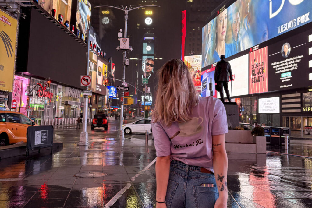 Kobieta tyłem w fioletowej koszulce DETOX z napisem Herbal Tea nocą na Times Square w Nowym Jorku